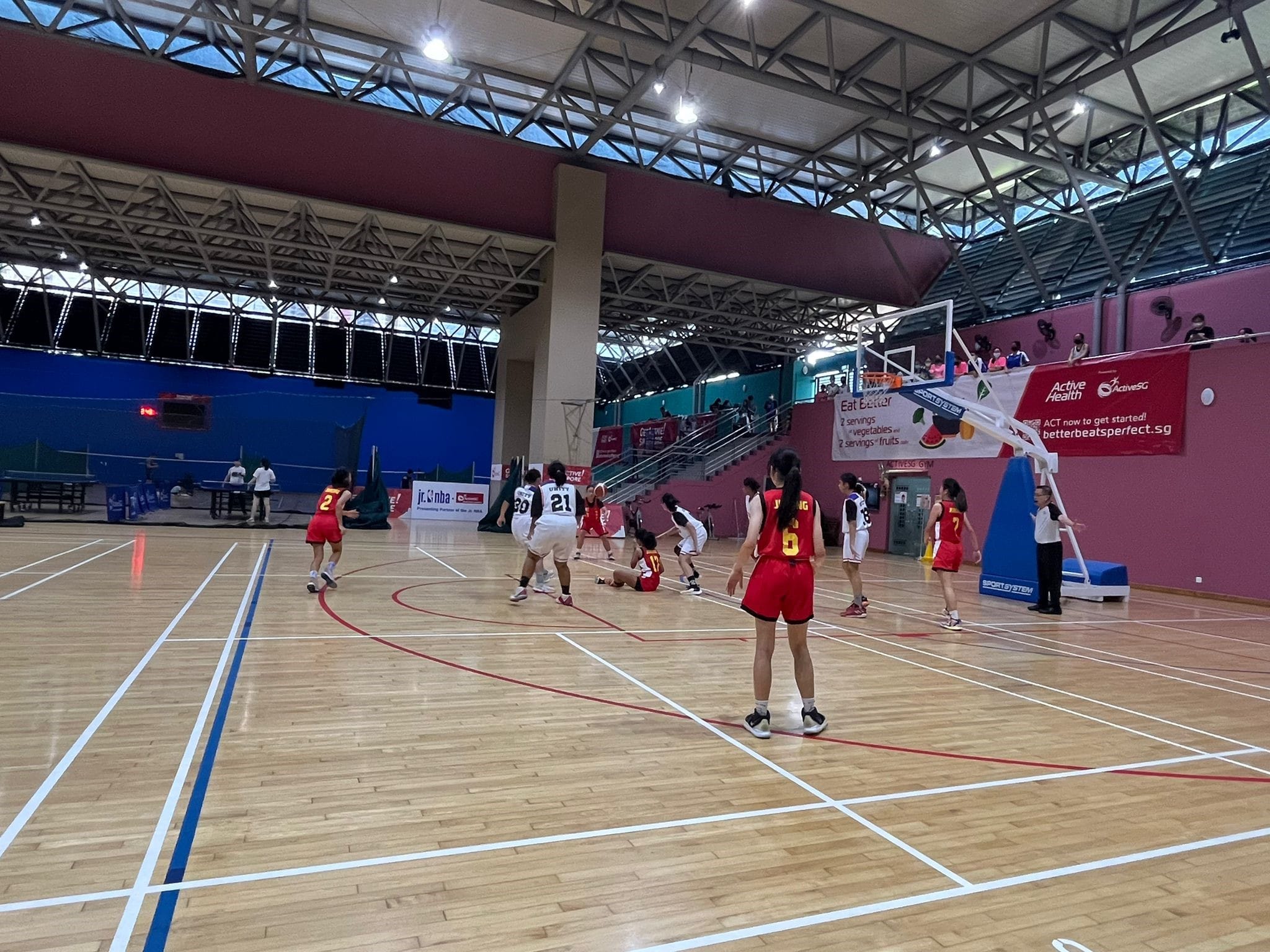 Girls in red and white uniforms play basketball on an indoor court near a hoop in a large covered arena.